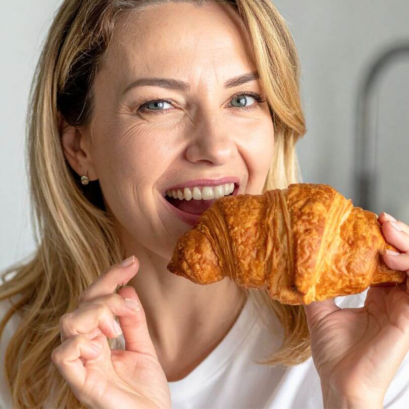 Donna sorridente che tiene in mano un fresco cornetto, assaporando il delizioso gusto dei 15 Cornetti proteici ripieni di crema al pistacchio.