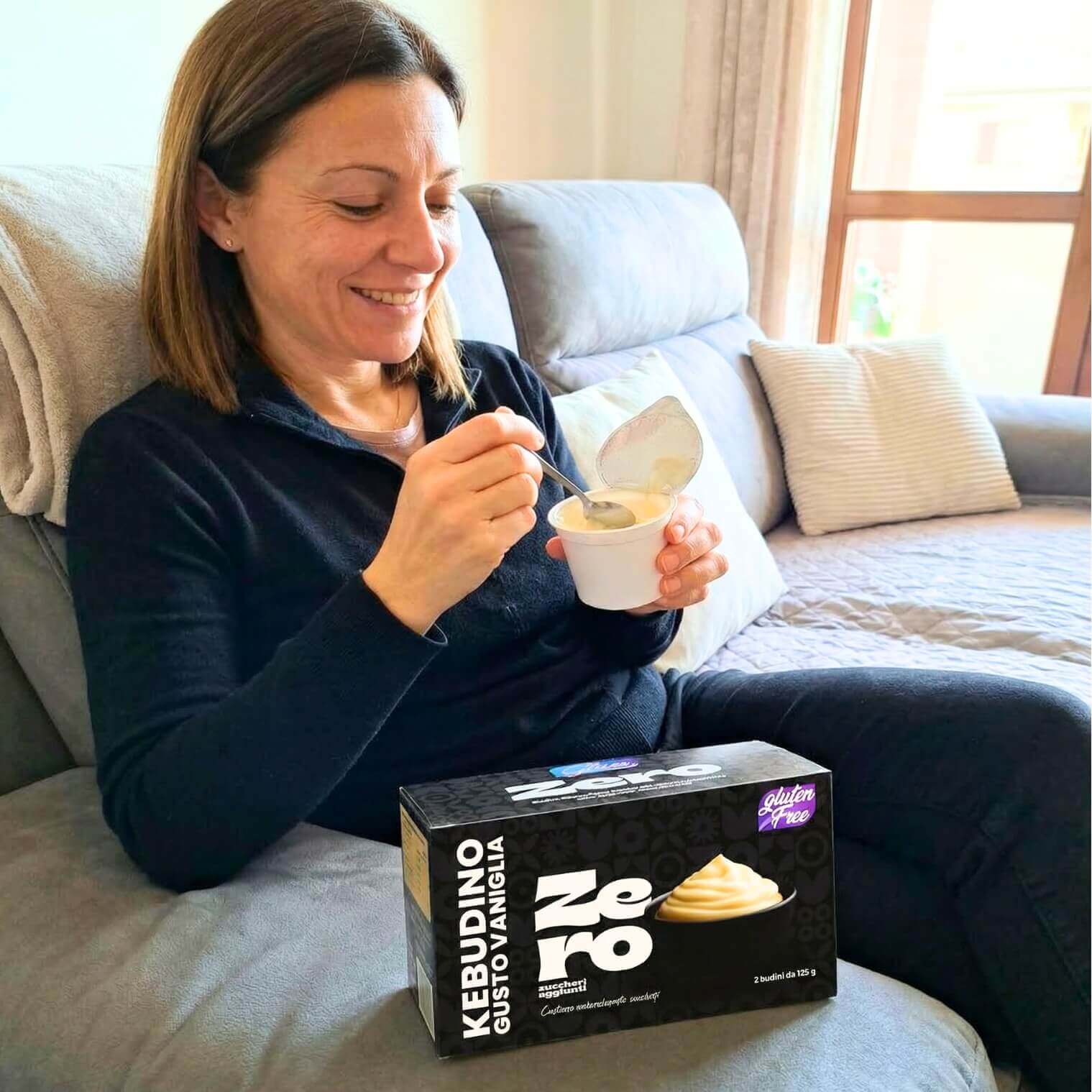 Donna che gusta Kebudino proteico alla Vaniglia su divano, con budino e confezione in vista.